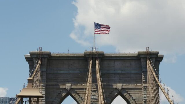 The US Flag waving in the wind on top of the Brooklyn Bridge on a sunny day with a blue sky and some clouds, static shot.
