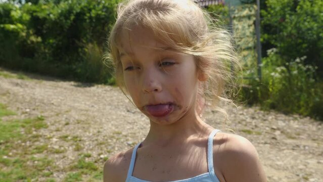A close-up of a blonde school-aged girl with a messy face makes faces while standing on the street. The child makes funny faces at the camera