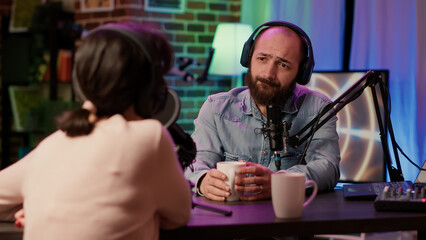 Man and woman having casual conversation and drinking coffe or tea in online podcast using professional recording equipment in home studio. Radio show host broadcasting interview with influencer.