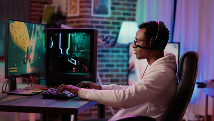 Gamer playing fast paced multiplayer space shooter competition using pc gaming setup having relaxing time at home. African american man streaming online simulation game while talking using headset. © DC Studio