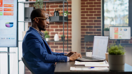 African american consultant working on business strategy with statistics report in office. Male employee using online research project to create startup presentation, writing email. Tripod shot.