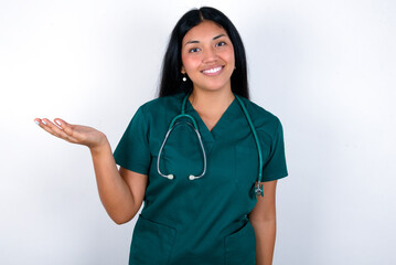 Doctor hispanic woman wearing surgeon uniform over white wall smiling cheerful presenting and pointing with palm of hand looking at the camera.