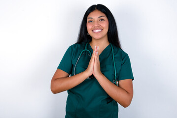 Doctor hispanic woman wearing surgeon uniform over white wall praying with hands together asking for forgiveness smiling confident.