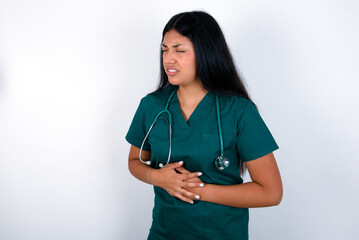 Doctor hispanic woman wearing surgeon uniform over white wall with hand on stomach because nausea, painful disease feeling unwell. Ache concept.
