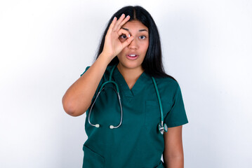 Doctor hispanic woman wearing surgeon uniform over white wall doing ok gesture shocked with surprised face, eye looking through fingers. Unbelieving expression.