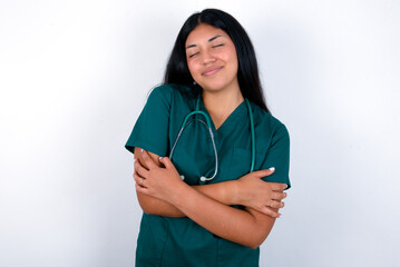 Doctor hispanic woman wearing surgeon uniform over white wall Hugging oneself happy and positive,...