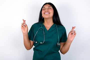 Doctor hispanic woman wearing surgeon uniform over white wall gesturing finger crossed smiling with hope and eyes closed. Luck and superstitious concept.