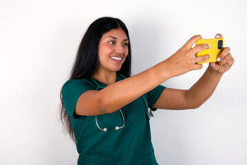 Doctor hispanic woman wearing surgeon uniform over white wall taking a selfie to post it on social...