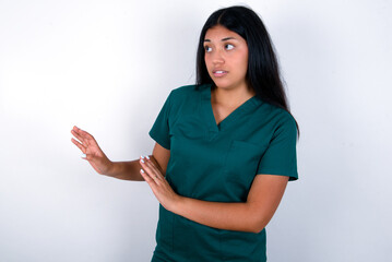 Displeased Doctor hispanic woman wearing surgeon uniform over white wall keeps hands towards empty...