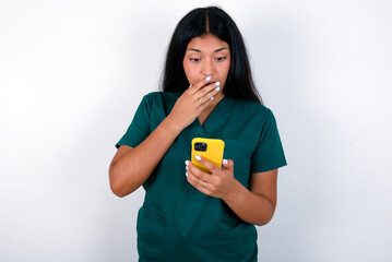 Doctor hispanic woman wearing surgeon uniform over white wall being deeply surprised, stares at smartphone display, reads shocking news on website, Omg, its horrible!