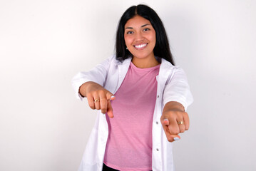 Young hispanic doctor girl wearing coat over white background cheerful and smiling poiting at camera
