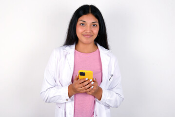 Portrait of serious confident Young hispanic doctor girl wearing coat over white background holding phone in two hands