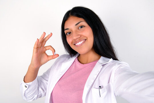 Portrait of pretty cheerful Young hispanic doctor girl wearing coat over white background make selfie show okey symbol