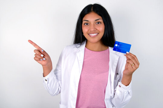 Smiling Young hispanic doctor girl wearing coat over white background showing debit card pointing finger empty space