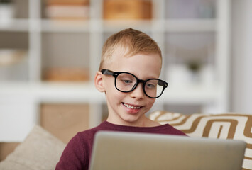 Smiling cute nerd boy in eyeglasses sitting on sofa and using laptop while searching for information