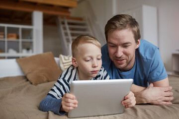 Serious blond boy lying on bed and using digital tablet while watching online movie with father