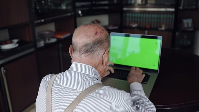 Intelligent grandfather working on laptop computer in home office, elderly man typing on trendy laptop keyboard and looking at green screen with tracking signs. Pensioner enjoying leisure activity