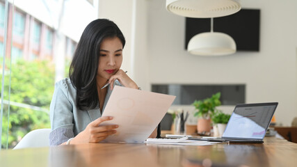 Serious young businesswoman reading statistics document, planning business strategy at office desk