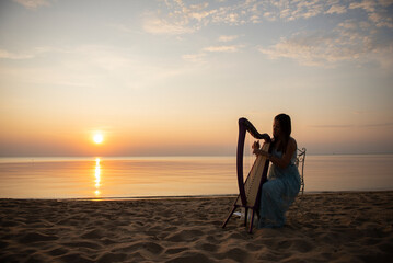 A girl in a flower dress plays on a Celtic harp by the sea at sunset