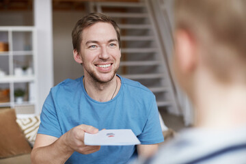 Portrait of smiling handsome young father with stubble giving birthday card to son while celebrating him at home