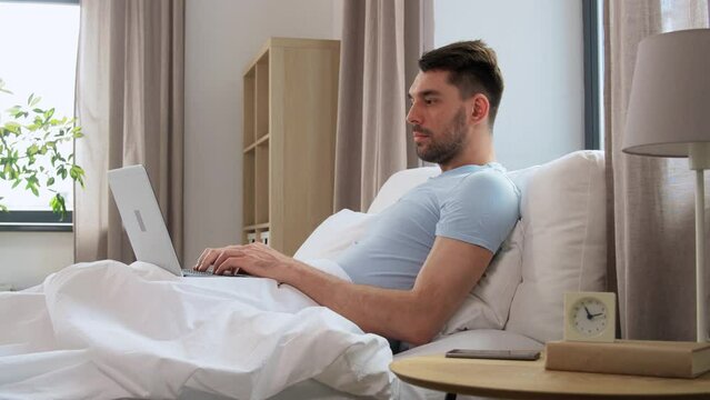 people, technology and remote job concept - man with laptop computer lying in bed at home bedroom