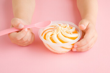 Little girl hand holding spoon and eating swirl white vanilla and yellow fruit ice cream on light pink table background. Pastel color. Closeup. Front view. Children cold sweet snack in plastic cup. - obrazy, fototapety, plakaty
