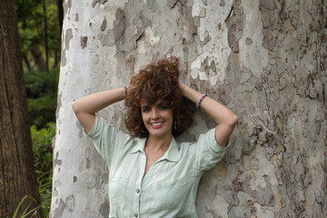 Portrait of attractive mature woman with curly brown hair and shirt, leaning against a tree trunk with her hands in her hair smiling happily. Concept happiness, hairstyle, curls, beauty.