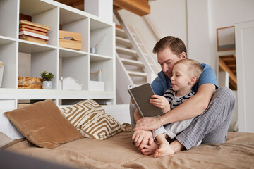 Smiling cute blond-haired son embraced by loving young father sitting on bed and reading book aloud