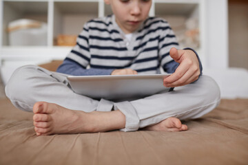 Close-up of curious barefoot child sitting with crossed legs on bed and scrolling on tablet