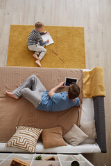 High angle view of young father lying on bed and surfing net on tablet while son sitting on carpet and drawing on paper