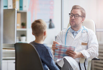 Fototapeta premium Content handsome young pediatrician with stethoscope around neck making notes in medical file while asking little boy about health complaints