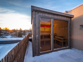 Small wooden finnish sauna with glass door on terrace of cottage.
