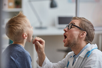 Expressive young pediatrician with stubble using Tongue depressor and asking boy to open mouth...
