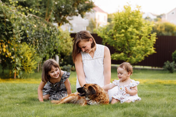 Family day, mother's day. Beautiful smiling young mom and two child daughters cuddling happy domestic dog on the backyard lawn. Idyllic family having fun with pet outdoors on summer holiday.