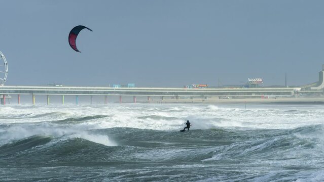 A dynamic footage of a kite surfer surfing under the harsh weather and going head-to-head against the big waves at the middle of the sea. There's a noticable amusement park in the background.