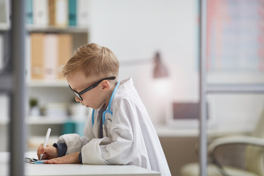 Serious concentrated little boy in doctors dress standing at table and making notes in medical file