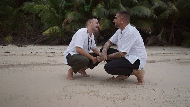  Happy Gay couple honeymooners in squatting position draw a heart shape on the sand and kiss on the beach of a tropical island 
