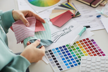 Close-up of unrecognizable female designer sitting at desk with papers and fashion sketches and choosing fabric for dress