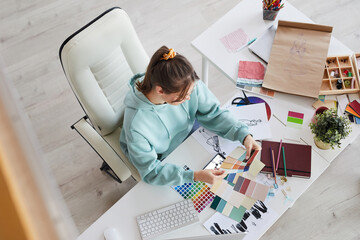 Above view of creative young designer sitting at table with massy sketches and choosing colors for...