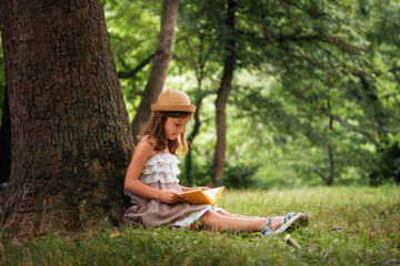 A cute schoolgirl girl in a straw hat and dress is sitting on the grass by a tree and reading a book. School holidays. The concept of independent extracurricular children's education