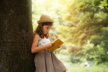Summertime. A beautiful schoolgirl girl in a straw hat and dress is standing by a tree and reading a book. School holidays. The concept of extracurricular children's education