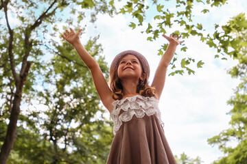 Portrait of a smiling Caucasian girl in a dress and a straw hat standing in a park and looking up at the crowns of trees. Summer holidays. The concept of a happy childhood and children's Day