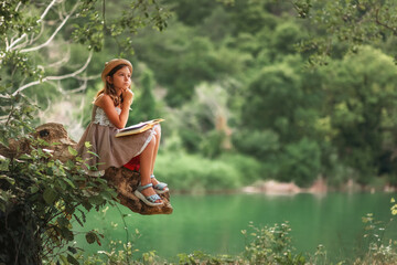 Pensive schoolgirl in a dress and a straw hat reading a book sitting on a old log. In the background are park and river. Copy space. The concept of children's education and school holidays