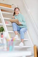 Smiling beautiful girl in glasses sitting on stairs with mug and looking into distance while drawing in sketchpad