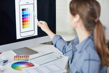 Rear view of female graphic designer sitting at table and pointing at computer monitor while...