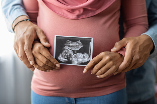 Pregnant Muslim Couple Standing Near Window And Holding Baby Sonography Photo