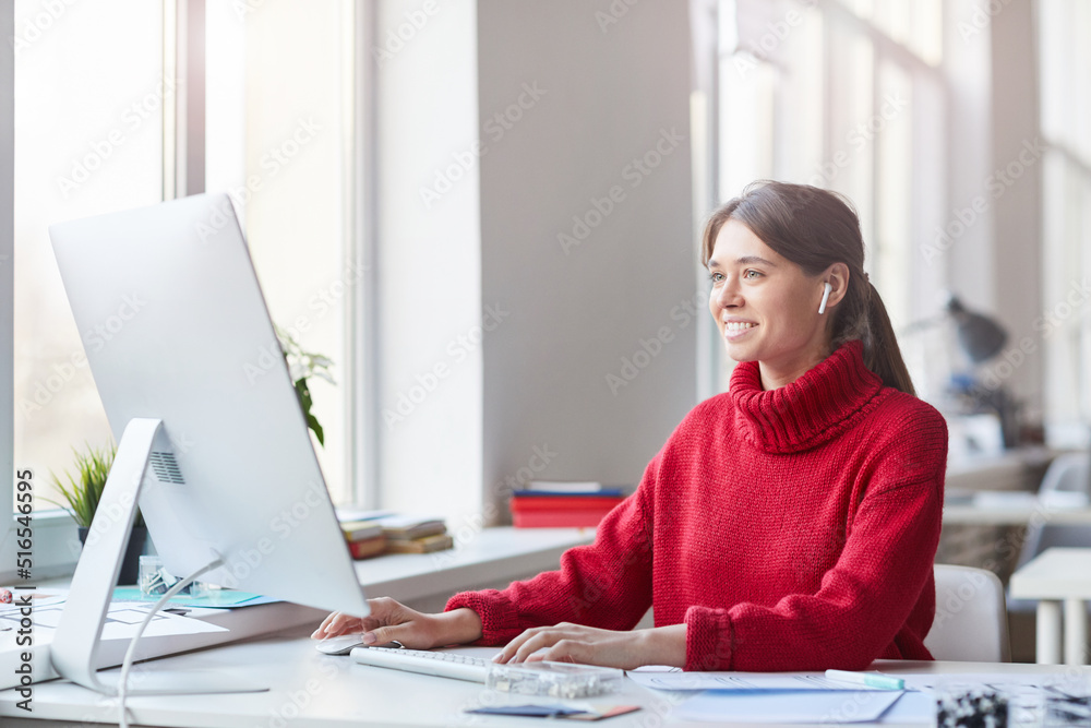 Wall mural Smiling pretty skilled young woman in red knitted sweater and wireless earbuds sitting at table in office and working with modern computer