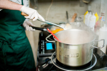 The cook in an apron and gloves is stirring the soup in a saucepan. Chef cooking a delicious dish in the kitchen. The owner of the soup kitchen or field kitchen interferes with the bucket in the soup. - obrazy, fototapety, plakaty