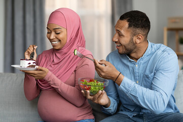 Black man offering salad to pregnant muslim wife while she eating cake
