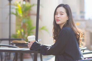 Asian woman with black suit sits on chair and holds coffee cup in her hands.